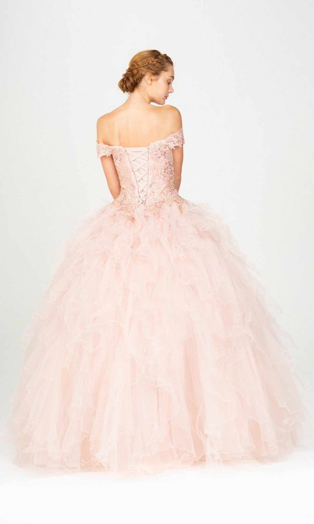 A woman faces away from the camera, wearing the Quinceanera Dress 9313 By Eureka—a blush pink gown with a lace-up corset and ruffled ball gown skirt.